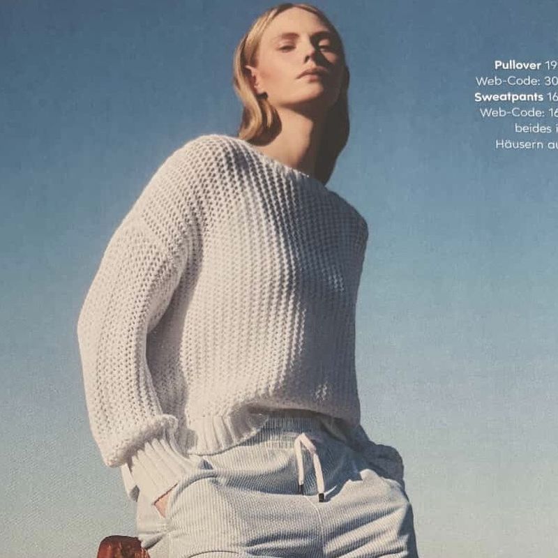 Adult female fashion model wears a white knit sweater and light sweatpants outdoors against a bright blue sky in soft, diffused light.