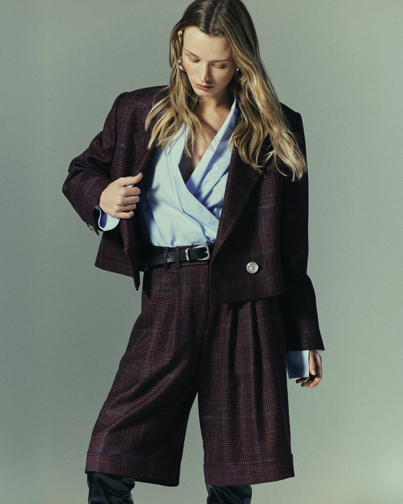 A blonde-haired woman poses indoors wearing a brown plaid blazer and matching culottes, accessorized with a black belt and gold earrings.