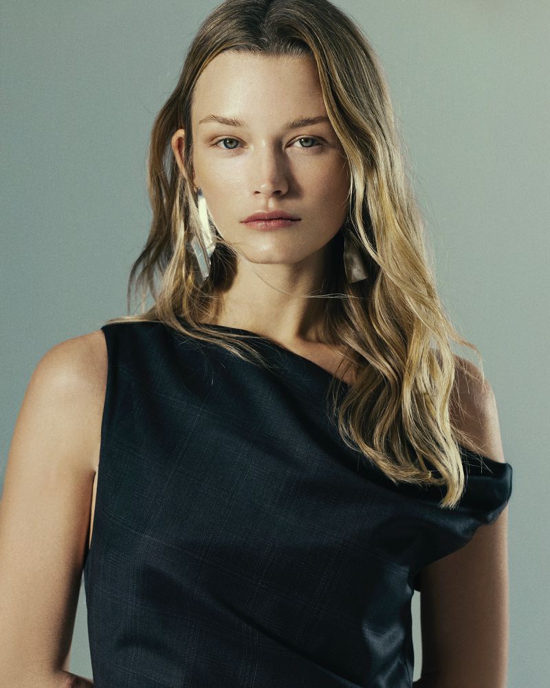Portrait of a blonde woman in a dark top with dramatic earrings, set against a pale grey background in a studio environment.
