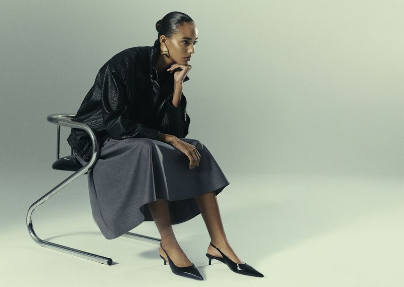 Elegant dark-skinned woman in black jacket and grey skirt sitting on a metal chair wearing pointed black slingbacks in studio lighting.