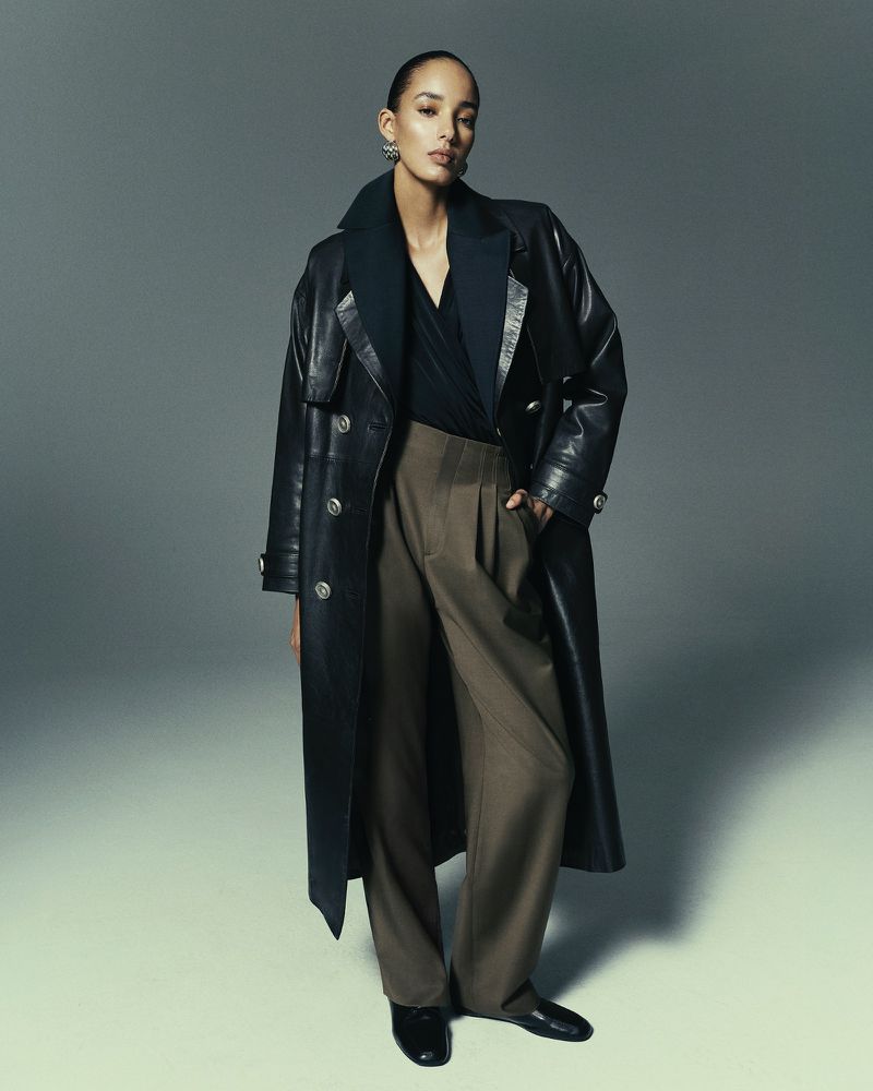Fashion shot of a woman in a long black leather coat and brown trousers posing in a photo studio against a grey background.