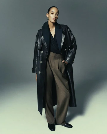 Fashion shot of a woman in a long black leather coat and brown trousers posing in a photo studio against a grey background.