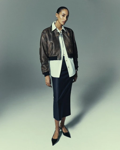 A woman in a brown bomber jacket, white shirt, and black skirt poses in a studio with a neutral background under bright, even lighting.