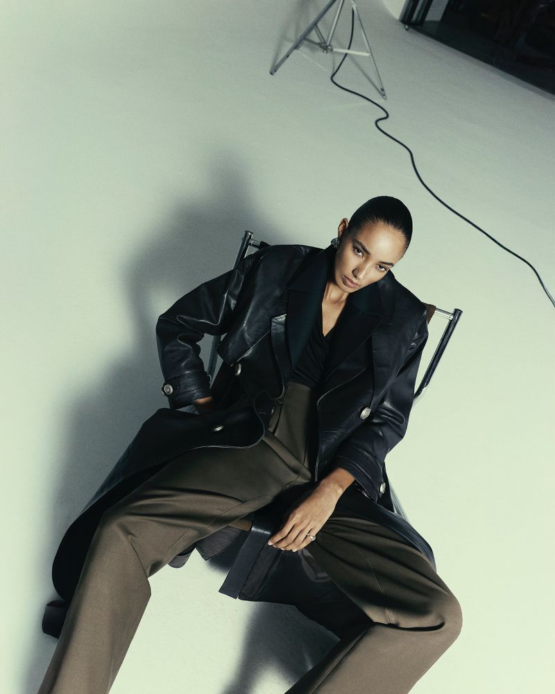 Model seated in a chair wearing a black leather coat and brown trousers against a white seamless background