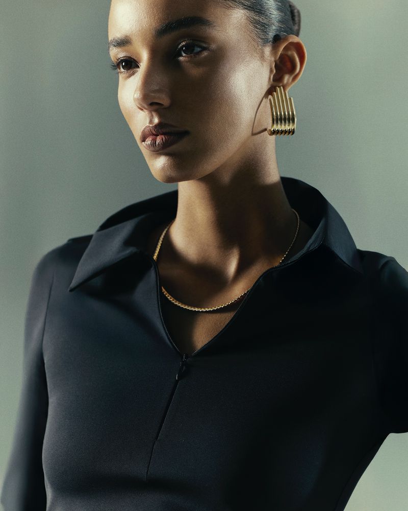 Portrait of a woman with fair skin wearing a black collared shirt and gold jewelry, including necklace and earrings.
