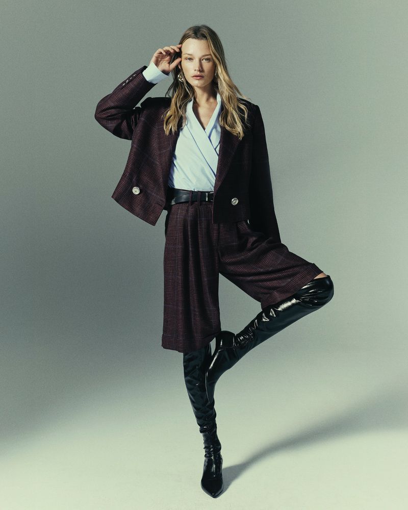 A fashionable woman in a maroon blazer and shorts, knee-high black boots, light blue blouse, and gold earrings poses in a studio.