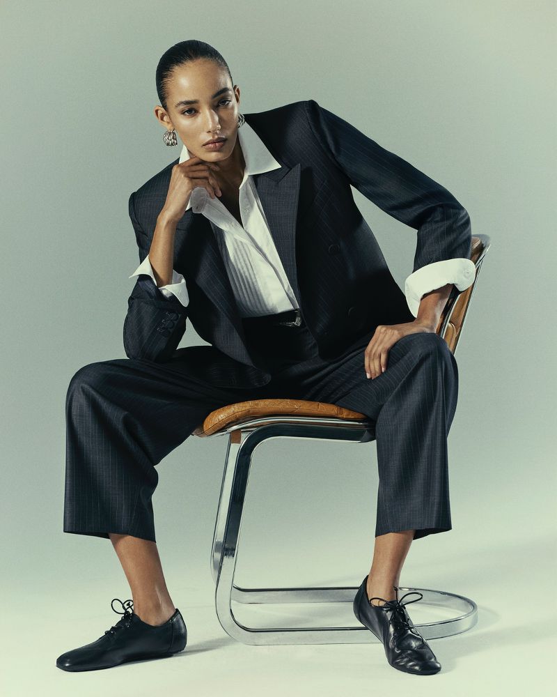 Elegant fashion portrait of a woman in a dark pinstripe suit, white blouse, and black shoes, seated on a tubular chair in a studio.