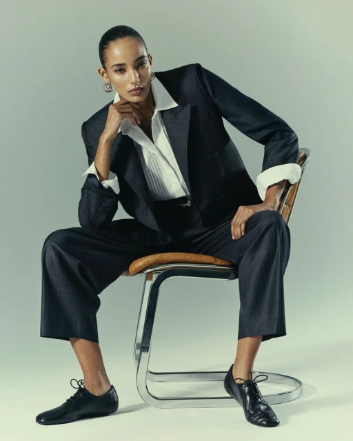 Elegant fashion portrait of a woman in a dark pinstripe suit, white blouse, and black shoes, seated on a tubular chair in a studio.