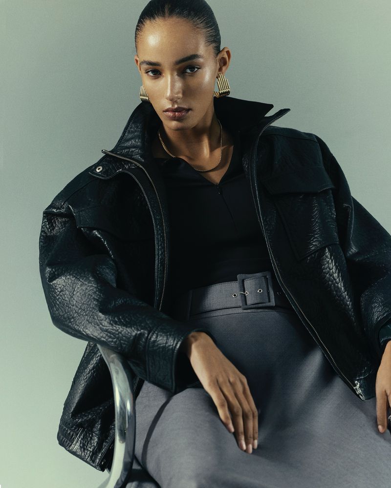 Portrait of an adult woman wearing a black jacket and gray skirt, seated in a studio with soft lighting.