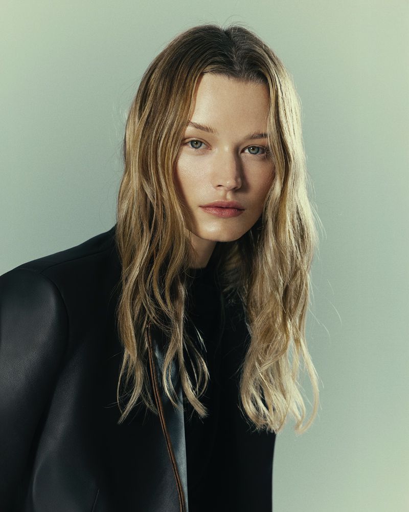 Portrait of a blonde woman in a black leather jacket against a light blue-grey background, showcasing a fashion or beauty aesthetic.