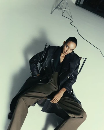 Model seated in a chair wearing a black leather coat and brown trousers against a white seamless background