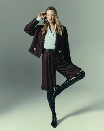 Model wearing a plaid blazer and shorts set with knee-high black boots against a neutral grey studio backdrop.