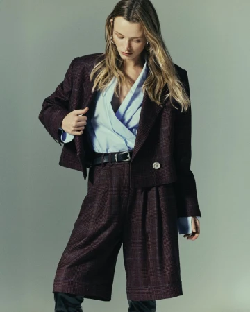 Model in plaid burgundy blazer and matching shorts with a light blue wrap top against a plain grey studio backdrop.