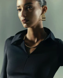 Close-up portrait of a woman wearing a black collared top, gold hoop earrings, and a delicate gold chain necklace against a grey studio background
