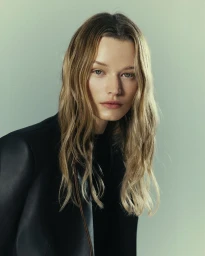 Portrait of a female model with blonde wavy hair wearing a black jacket against a plain light green background