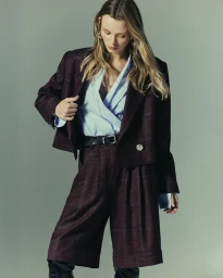 Model in plaid burgundy blazer and matching shorts with a light blue wrap top against a plain grey studio backdrop.
