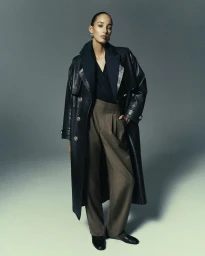 Model in black leather trench coat and brown trousers posing against a grey studio backdrop for Acaria Studio.