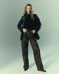 Model standing against a soft neutral studio backdrop wearing a dark leather blazer over a black top and brown leather trousers