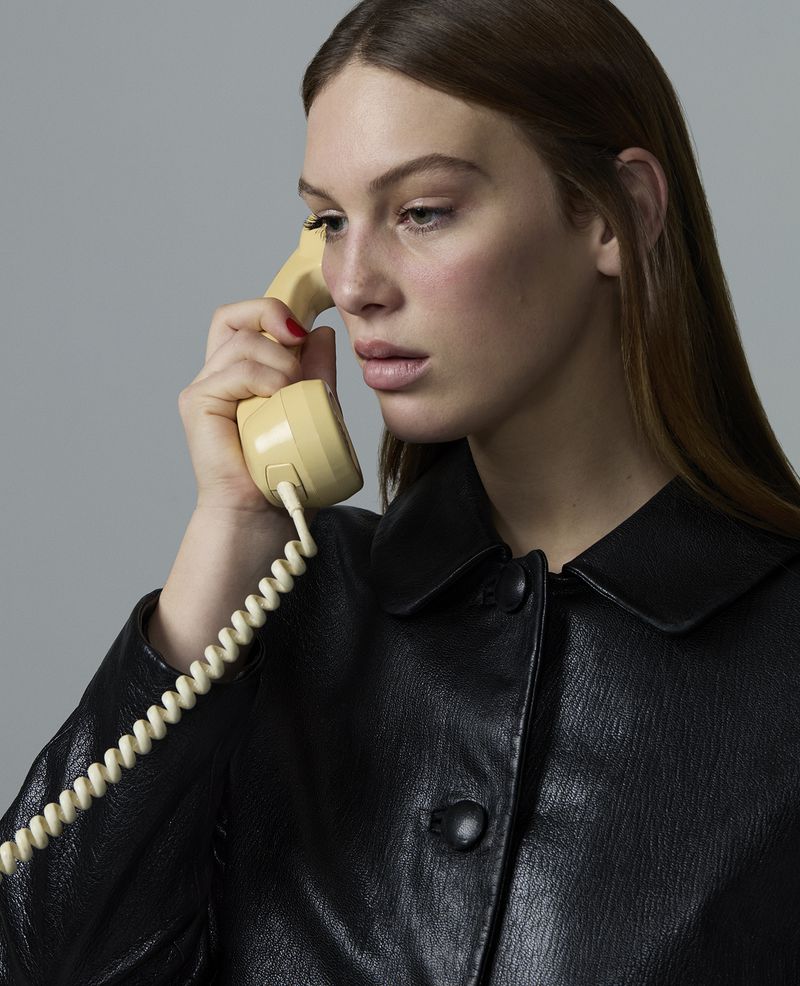 Portrait of a young woman in a black leather jacket holding a vintage rotary phone, set against a neutral grey backdrop in a studio.
