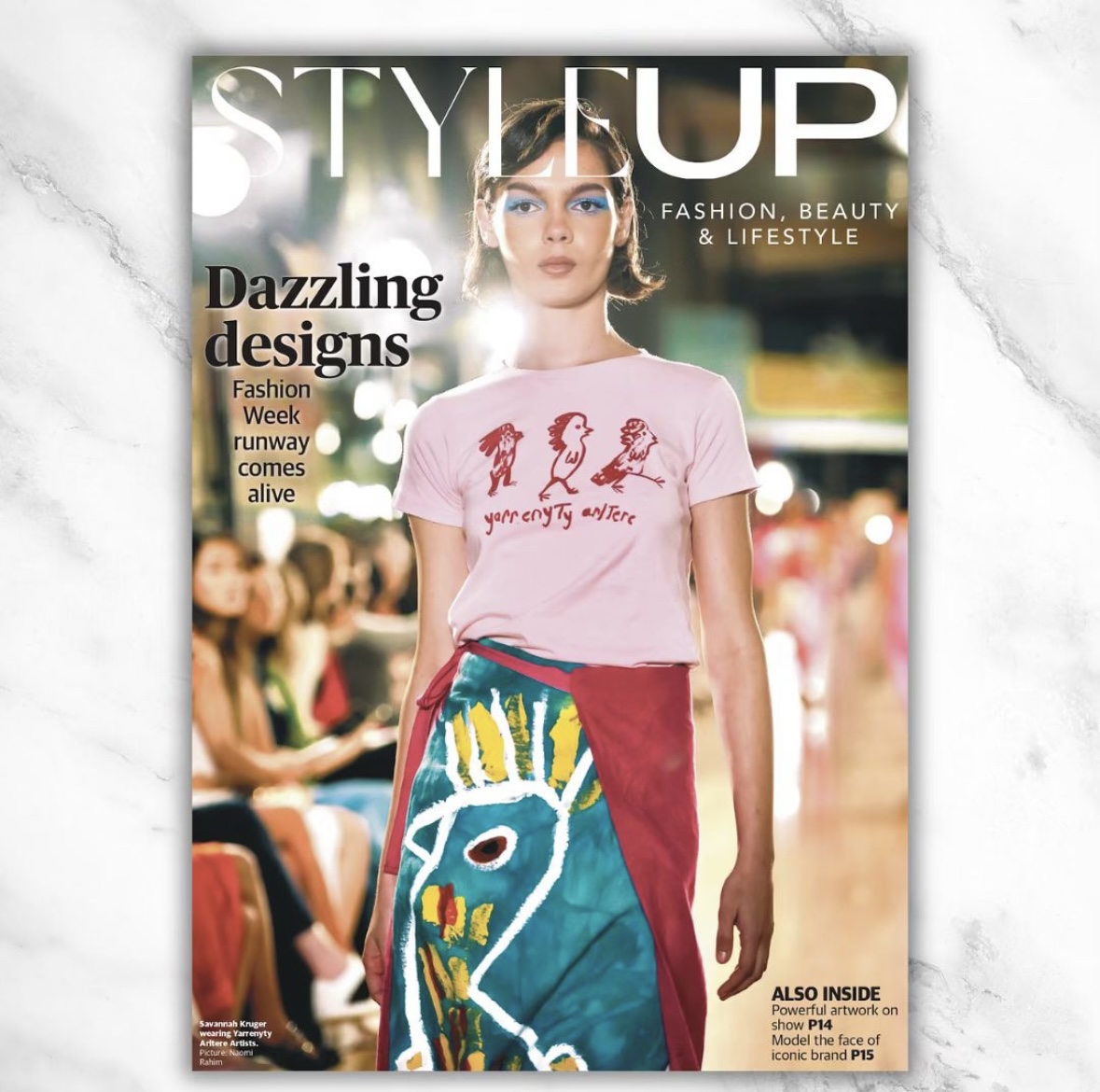 Magazine cover featuring a model on a runway wearing a t-shirt with graphics and text, plus a brightly colored apron skirt.