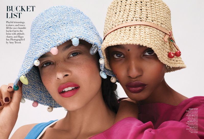 Two women wearing straw bucket hats in a fashion shot. One has cherry adornments, and the other has pastel-colored ball adornments.