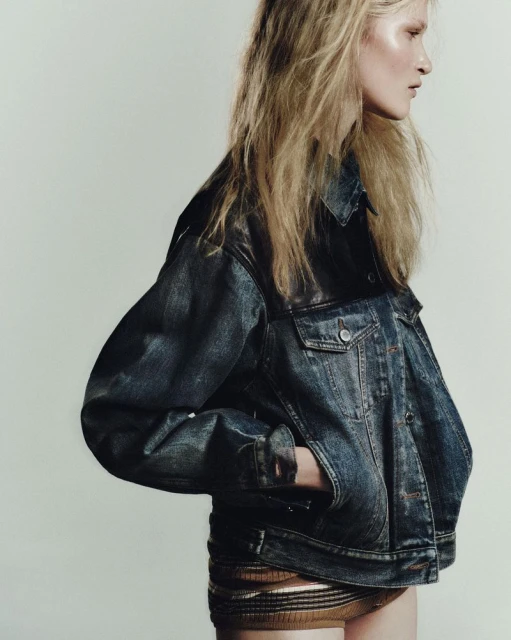 A blond woman in a denim jacket with a hand in her pocket poses against a white background in a fashion shot.