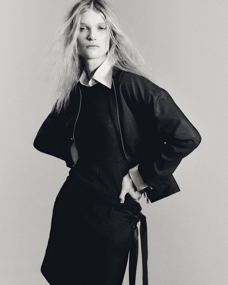 Monochrome studio portrait of a slender adult woman posing with hands on her hips wearing a black sweater dress and bomber jacket.