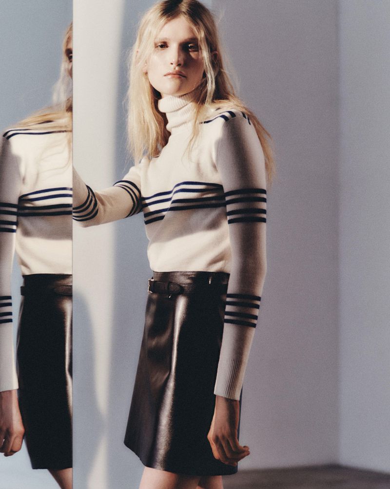 Fashionable blonde model in a striped turtleneck and black leather mini skirt poses in front of a reflective surface in a studio.