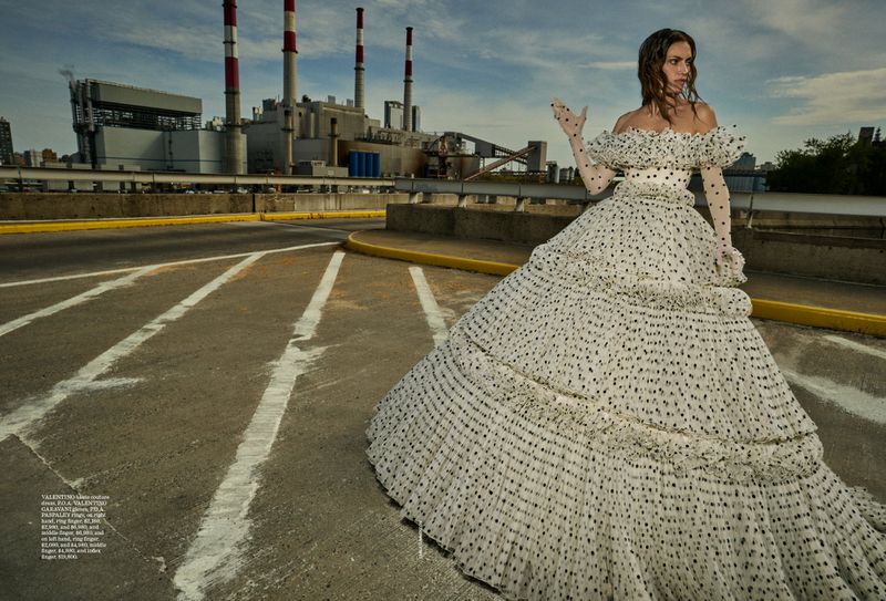 Adult female model in a polka-dot gown and gloves poses on an asphalt road with an industrial complex in the background.