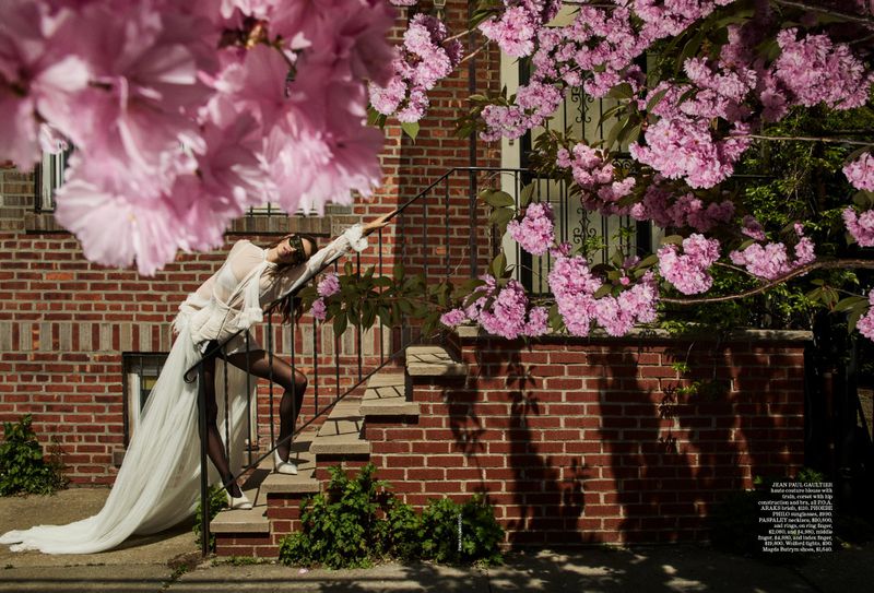 A woman posing in white on steps near a brick building surrounded by flowering trees, bathed in soft light, showcasing fashion outdoors.