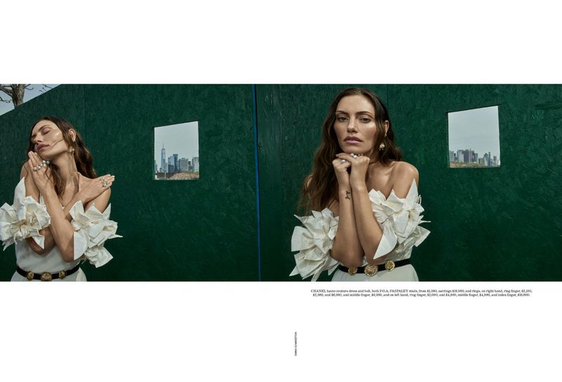 Fashion shot of woman in off-shoulder white dress and gold belt, posing in front of a green wall with a city skyline reflection.