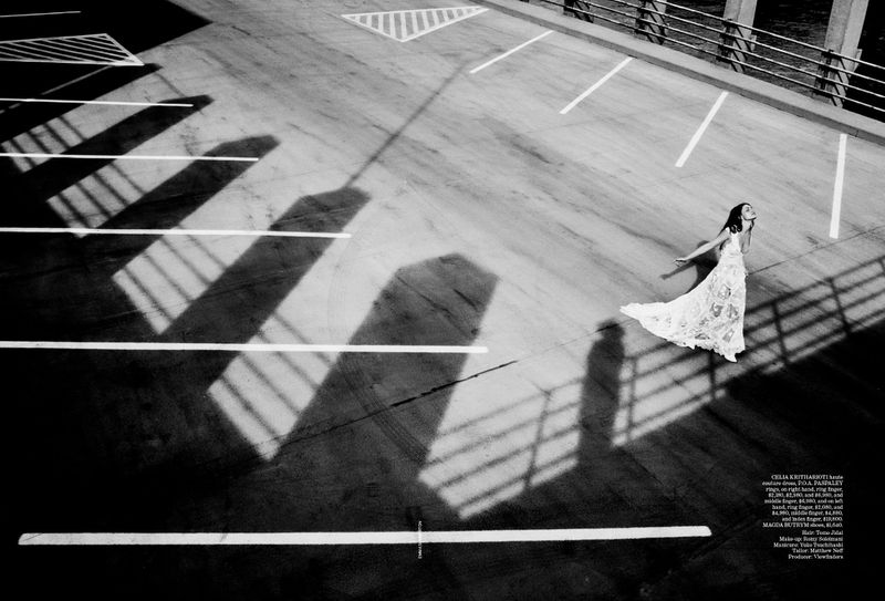 Woman in white lace dress stands on concrete parking lot surface with long shadow patterns from bridge or building in black and white.