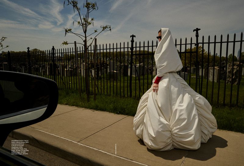 Woman in oversized white dress with hood stands on sidewalk near black metal fence and road on sunny day with light clouds.