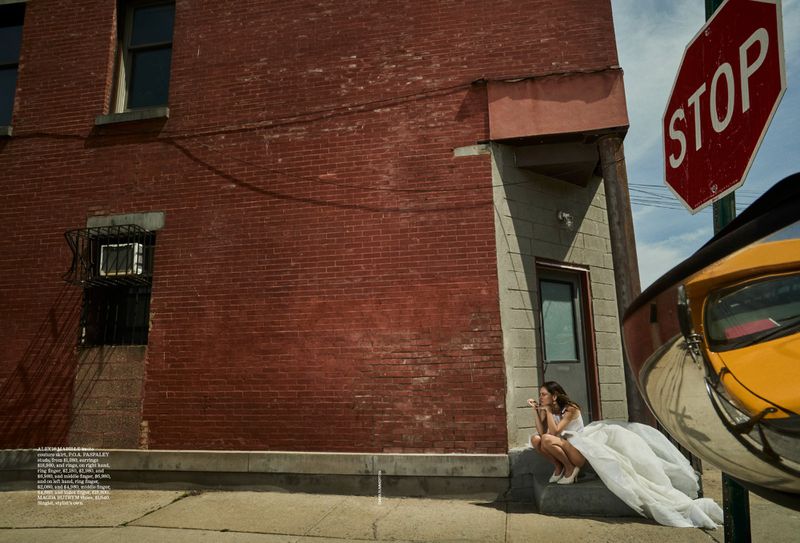 Woman in white dress sits on sidewalk steps near a red brick building and a stop sign with a yellow vehicle reflected.