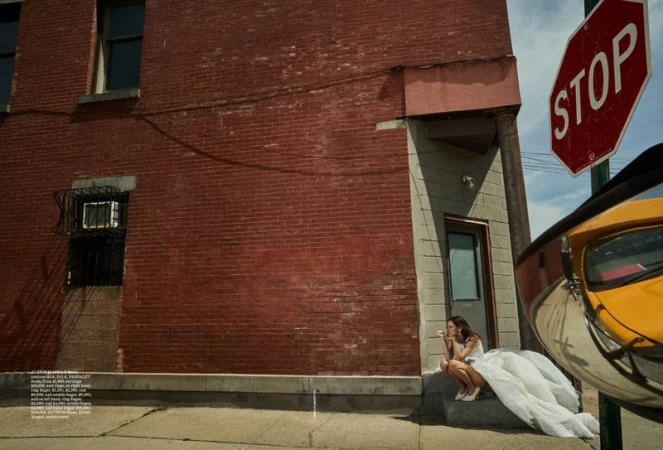 Woman in white dress sits on sidewalk steps near a red brick building and a stop sign with a yellow vehicle reflected.