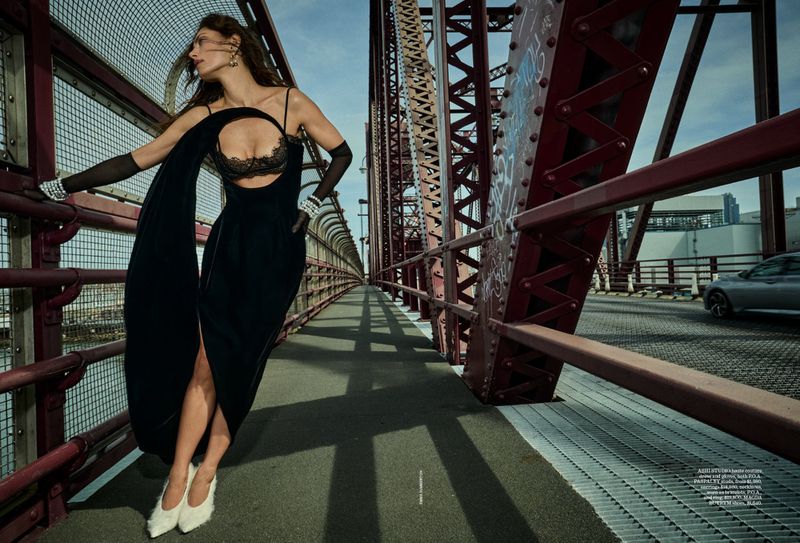 Fashion model in black evening gown on a bridge with a city background, wearing long gloves and pointed-toe white heels.