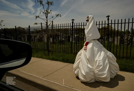 Phoebe Tonkin wearing a voluminous white hooded gown walking on a sidewalk beside a black iron fence.