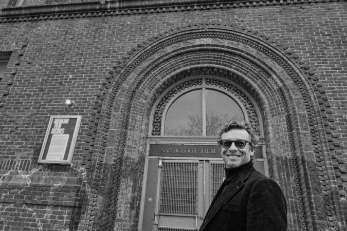 Black and white portrait of smiling adult male in front of brick building with arched doorway and signage in an urban environment.
