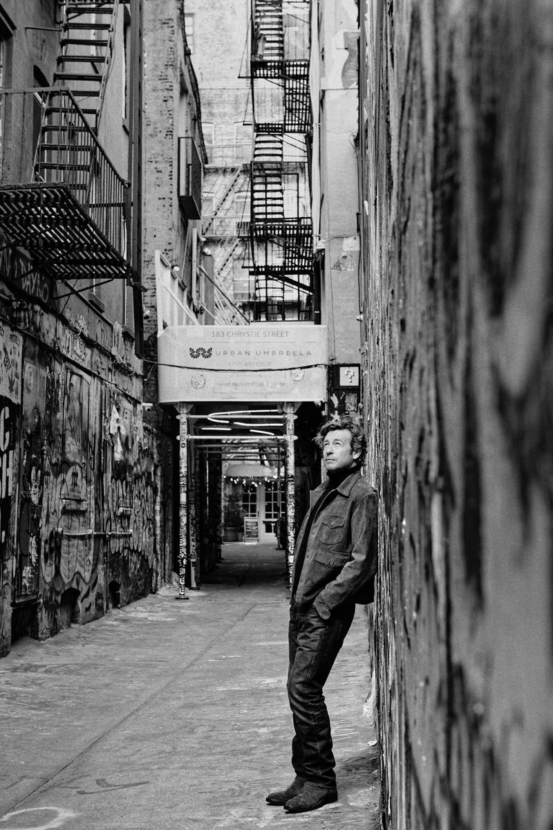 Monochrome portrait of an adult male leaning against a graffiti-covered wall in an urban alleyway with fire escapes and building scaffolding.