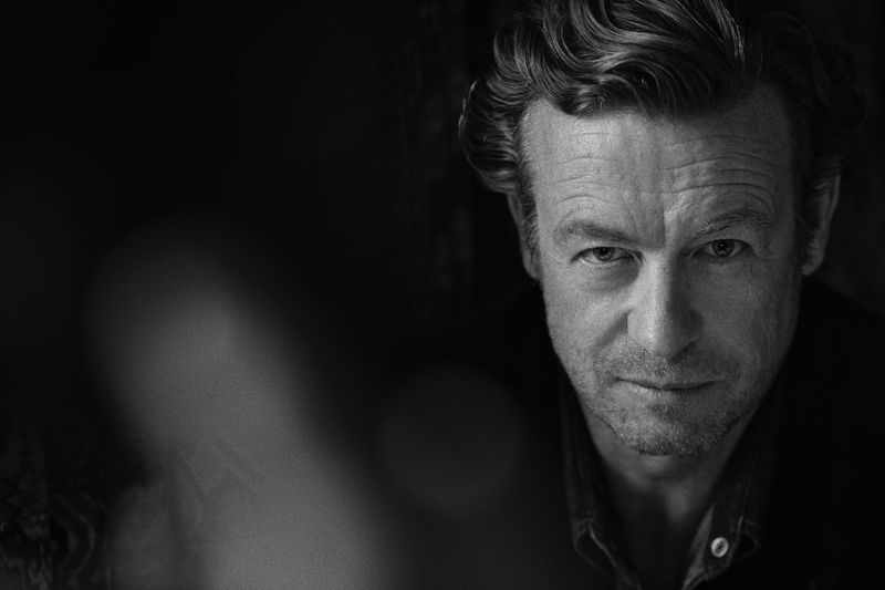 Monochrome portrait of an adult male with wavy hair, looking directly at the camera with a slight smirk, set against a dark background.