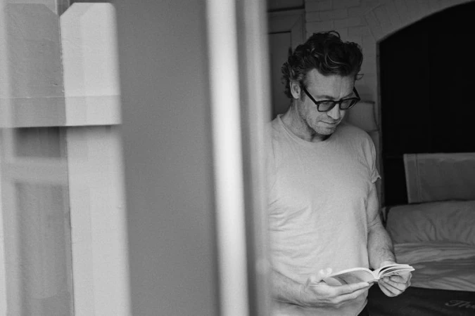 Black and white portrait of a man with glasses, reading a book indoors, near a bed. Natural light illuminates the scene.
