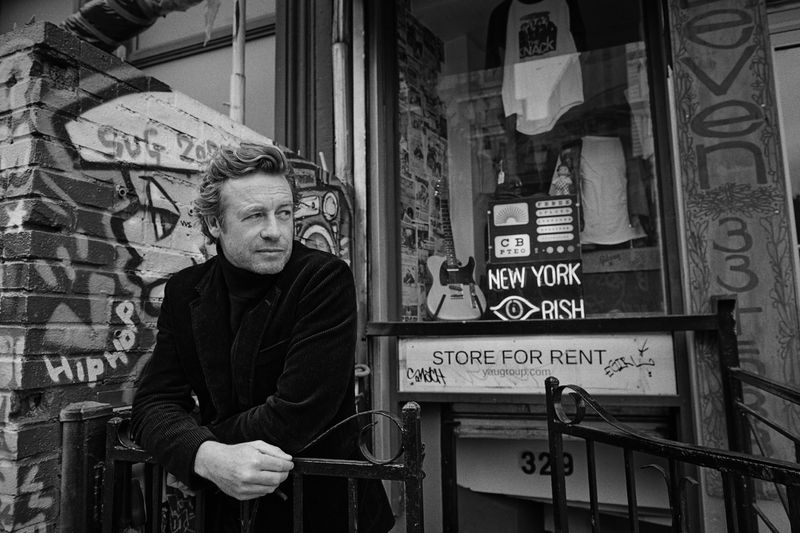 Black and white portrait of a man with fair hair leaning on a metal fence in front of a building with graffiti and a store window.