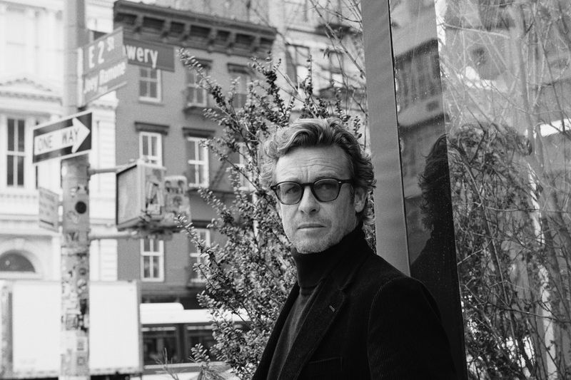 Black-and-white portrait of Simon Baker outdoors, with buildings, foliage, and street signs in the background, captured at eye level.