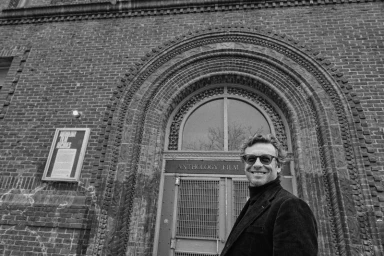 Actor Simon Baker wearing dark sunglasses and a dark blazer standing outside an arched brick building entrance.