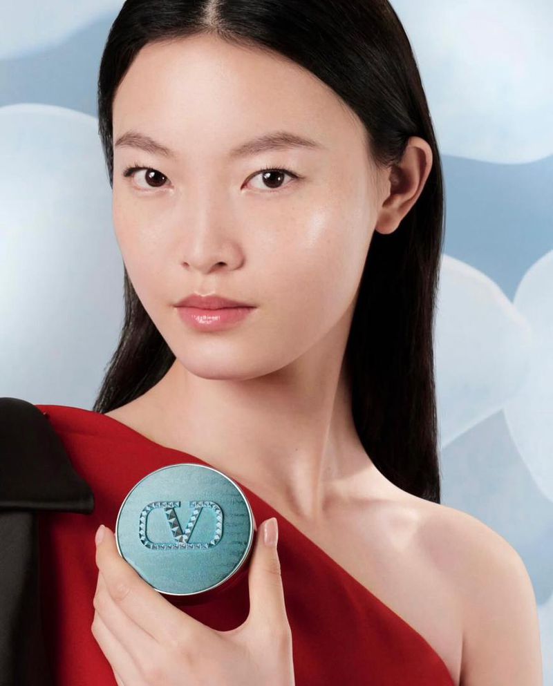 Close-up of an Asian woman with black hair holding a light blue Valentino beauty compact, wearing a red dress against a light blue background.