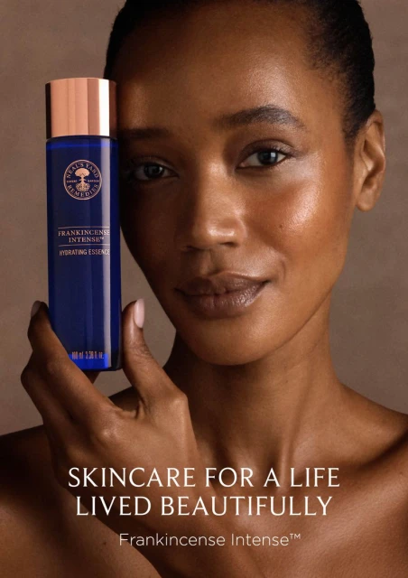 An African-American woman poses next to a blue glass bottle of skincare product against a neutral background in a beauty advertisement.