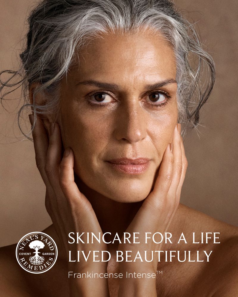 Close-up portrait of an older woman with silver-streaked hair holding her face with both hands against a neutral warm background.