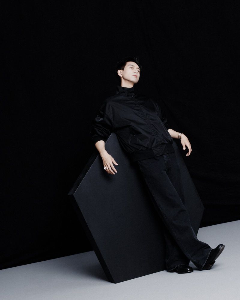 Portrait of a young adult male in an all-black outfit, leaning against a black cube in a photo studio with dark and light contrast.