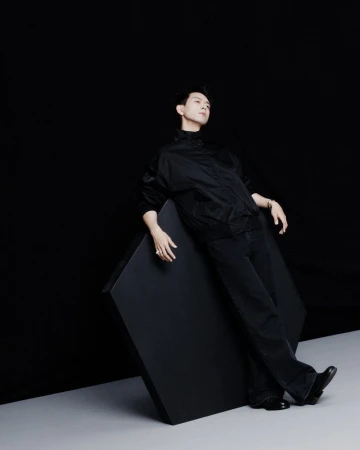 Portrait of a young adult male in an all-black outfit, leaning against a black cube in a photo studio with dark and light contrast.