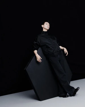 Actor Li Xian wearing a black jacket and pants leaning against a large dark geometric prop on a two-tone studio set.